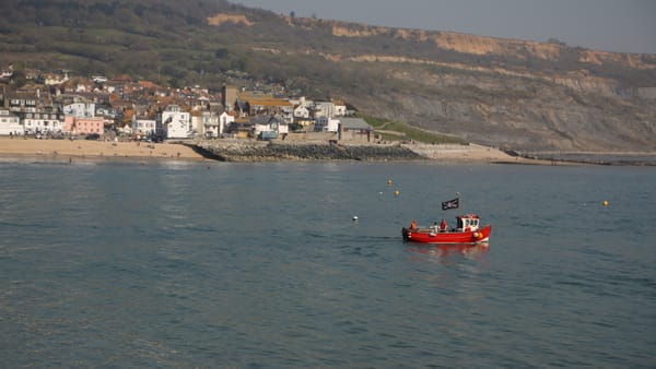 A red fishig boat is to the right of the viewer as we look back over the sea to the coast line with a small town and cliffs in the background.